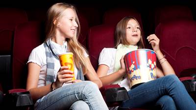 To piger sidder ved siden af hinanden på biografsæder. Den ene spiser popcorn.
