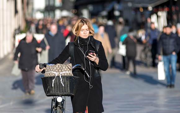 Kvinde trækker sin cykel mens hun kigger ned på sin smartphone med høretelefoner. Hun går på Strøget